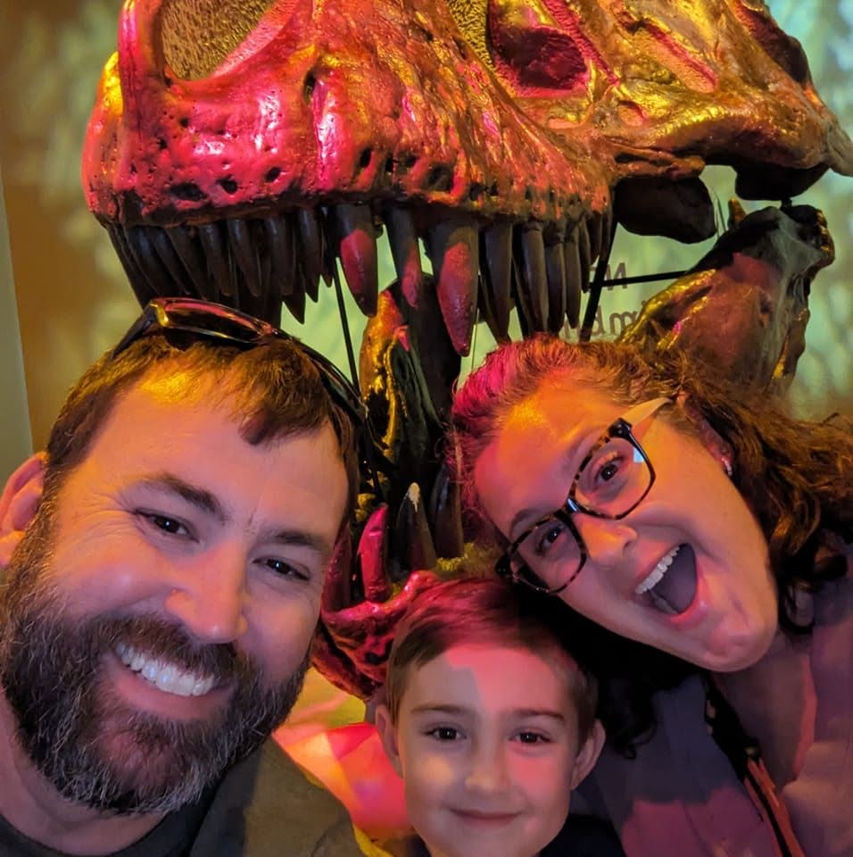 Matthew laughing with his wife Jenn and son Miles in front of a dinosaur skull on one of their family adventures